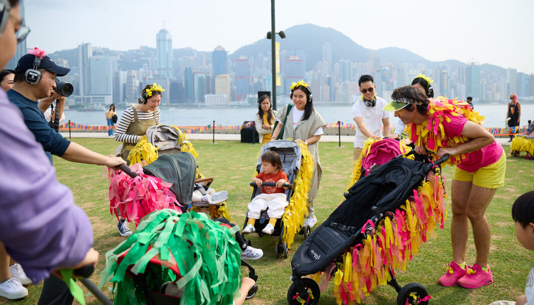 Pram People (Hong Kong Edition) production photo: West Kowloon Cultural District Authority. Parents wearing headphones and pushing small children in prams decorated with colourful ribbons, and a Polyglot artist in a bright pink and yellow costume with a pram, form a circle. They are on a green lawn next to a harbour, with a city skyline and a mountain range in the background.
