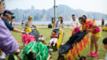 A Pram People production photo. Performers dressed in bright colours push black prams decorated with colourful streamers with adults who also push children in prams. They congregate on a green lawn. In the background is a lake. On the other side of the lake are buildings and hills. Photo courtesy of West Kowloon Cultural District Authority.