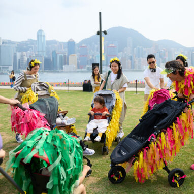 Pram People (Hong Kong Edition) production photo: West Kowloon Cultural District Authority. Parents wearing headphones and pushing small children in prams decorated with colourful ribbons, and a Polyglot artist in a bright pink and yellow costume with a pram, form a circle. They are on a green lawn next to a harbour, with a city skyline and a mountain range in the background.