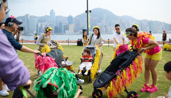 A Pram People production photo. Performers dressed in bright colours push black prams decorated with colourful streamers with adults who also push children in prams. They congregate on a green lawn. In the background is a lake. On the other side of the lake are buildings and hills. Photo courtesy of West Kowloon Cultural District Authority.