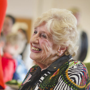 A photograph of Naomi Tippett AM. She is sitting in a chair at a Polyglot Theatre 40th birthday event and smiling. Photographer: Theresa Harrison, 2018