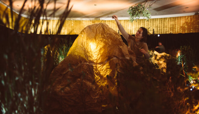 A When the World Turns production photo. A Polyglot artist brushes a branch across a large 'tent' of wrinkled brown paper that is illuminated from within. They are in a darkened space filled with plants. Photographer: Theresa Harrison