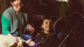 A When the World Turns production photo. A child in a wheelchair looks up at someone out of frame. An adult sits next to them, offering them a crumpled ball of brown paper. They are in a darkened space surrounded by plants. Photographer: Theresa Harrison