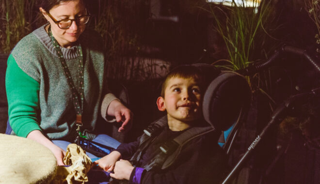 A When the World Turns production photo. A child in a wheelchair looks up at someone out of frame. An adult sits next to them, offering them a crumpled ball of brown paper. They are in a darkened space surrounded by plants. Photographer: Theresa Harrison