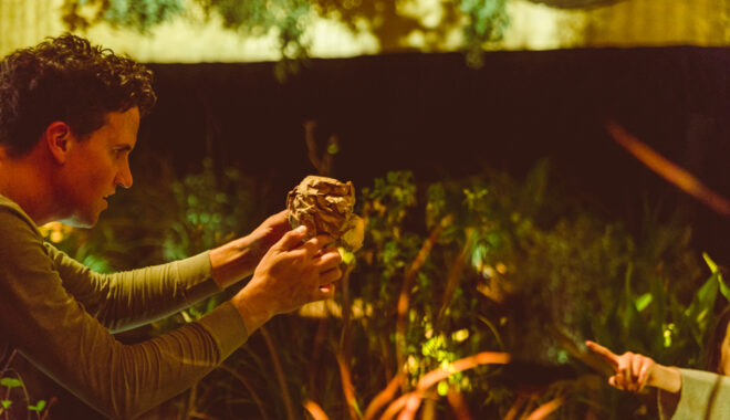 A When the World Turns production photo. A Polyglot artist holds up a crumpled ball of brown paper. A child's arm can be seen pointing towards it. They are in a theatrically lit space filled with plants. Photographer: Theresa Harrison