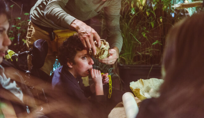 A When the World Turns production photo. A Polyglot artist holds a crumpled ball of brown paper near a child in a wheelchair. They are in a theatrically lit space filled with plants. Photographer: Theresa Harrison