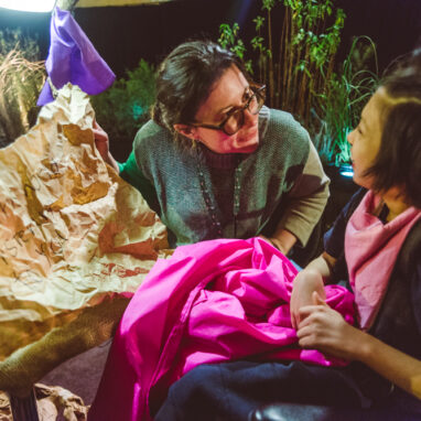 A When the World Turns production photo. An adult shows a message written on wrinkled brown paper to a child, who is smiling. They are surrounded by green plants. Photographer: Theresa Harrison