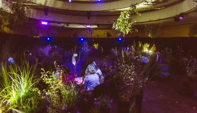 A When the World Turns production photo. A large indoor space, divided into circles with plants, theatrically lit. People sit at small tables in the centre of each circle. Photographer: Theresa Harrison