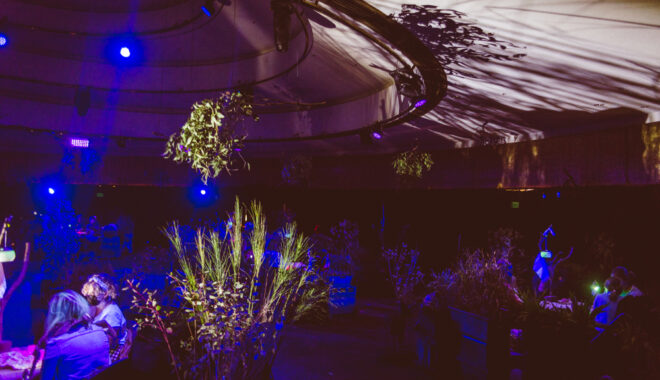 A When the World Turns production photo. A large dark, indoor space, divided into circles with plants, theatrically lit. People sit at small tables in the centre of each circle. The plants cast dramatic shadows on the ceiling. Photographer: Theresa Harrison
