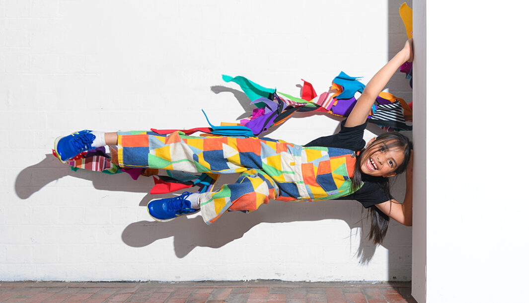 A Come Back Home photo. A child in colourful overalls is suspended sideways against white walls. A long piece of colourful bunting is cascading onto them. They are smiling at the camera. Photographer: Sarah Walker