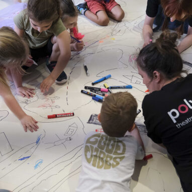 A Family Forts production photo. Two Polyglot artists and a group of small children draw together on a large white surface on the floor. Photographer: Suzanne Phoenix