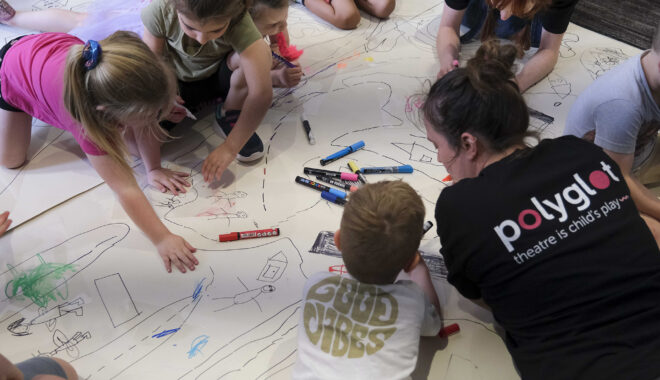 A Family Forts production photo. Two Polyglot artists and a group of small children draw together on a large white surface on the floor. Photographer: Suzanne Phoenix