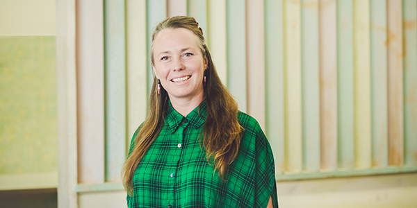 IMAGE: An indoor portrait of Cat Sewell, standing in front of a pastel-coloured heritage partition. She wears a green checked shirt, her long hair over her shoulders, and she smiles at the camera. Photographer: Theresa Harrison