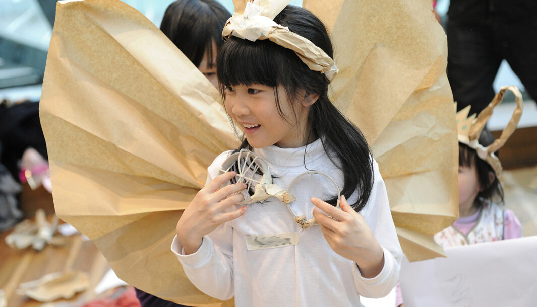 A Paper Planet production photo. A child in a white shirt with long dark hair wears large handmade brown paper wings and a handmade paper crown. They are smiling at something out of frame. Photographer: Martin Reddy, 2011