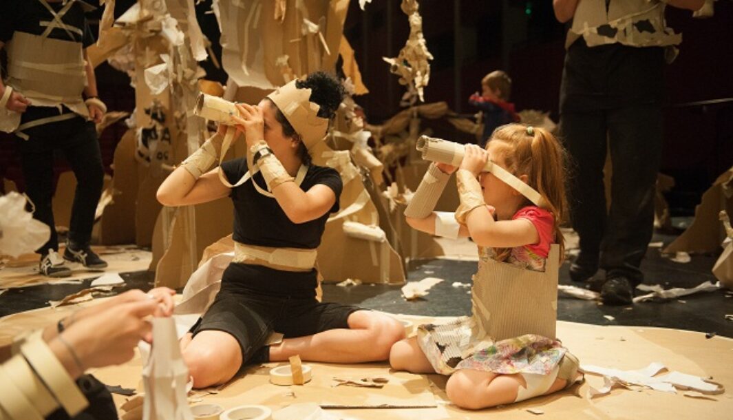 A Paper Planet production photo. A Polyglot performer and a child sit in a forest of tall brown cardboard trees, adorned with paper plants and creatures. They look around with paper binoculars. Other performers, children and families are visible amongst the trees, creating and playing. Photographer: Sarah Walker