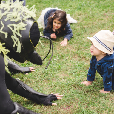 A Bees production photo. A bee in costume is kneeling towards the ground with both hands placed on grass. Two children, one wearing a hat, look up at the bee in awe.