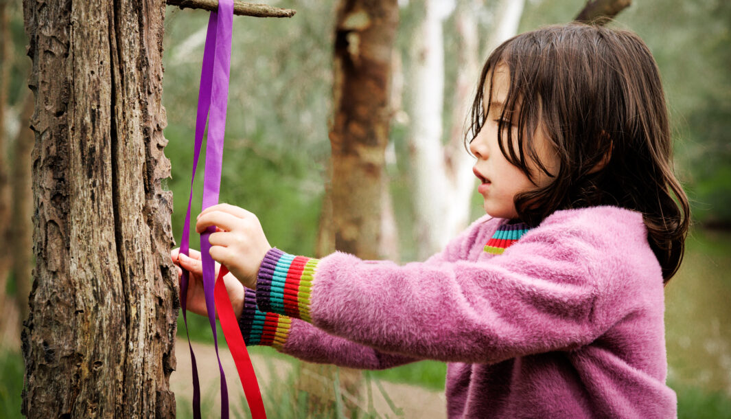 A Forest promo photo. A child in a pink jumper creates and plays with colourful ribbons on a tree branch. Photographer: Darren Gill