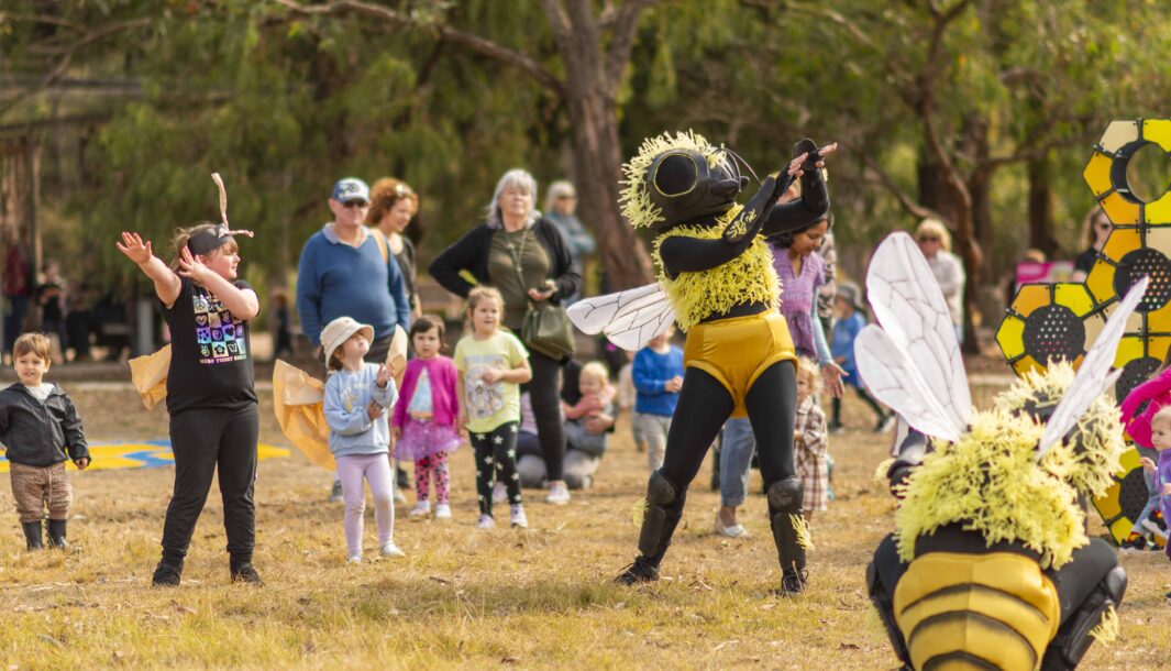 Bees production photo. Royal Botanic Gardens Cranbourne. A Polyglot artist in a Bee costume demonstrates a dance movement which a child mirrors to the right of them. There's an another Bee crouched to the ground. There are families in the background.