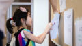 A Come Back Home production photo. A young child wearing a backpack with their hair tied in a ponytail. The child attaches paper with a drawing on it to a brown wall, with the help of an adult. Photo: Studio Znke, courtesy of Esplanade - Theatres on the Bay, Singapore
