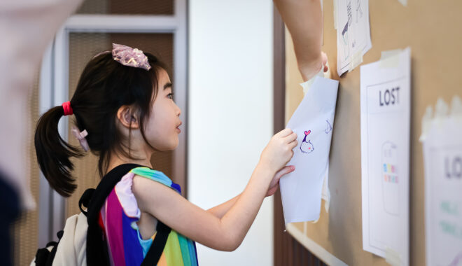 A Come Back Home production photo. A young child wearing a backpack with their hair tied in a ponytail. The child attaches paper with a drawing on it to a brown wall, with the help of an adult. Photo: Studio Znke, courtesy of Esplanade - Theatres on the Bay, Singapore