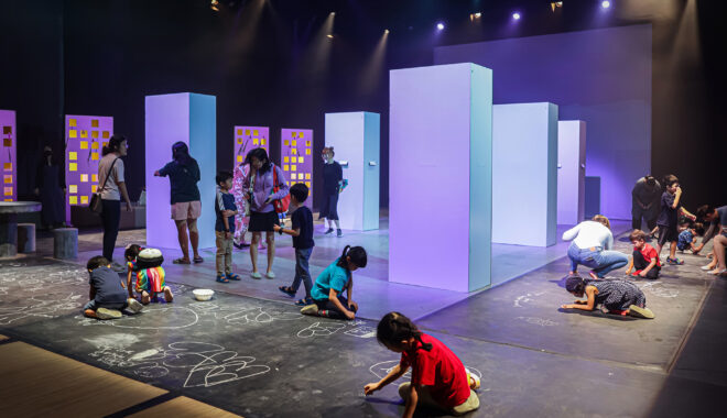 A Come Back Home production photo. Children draw on a floor using chalk under theatre lighting. Behind them, children and adults stand and talk on a stage decorated with tall cardboard buildings. Photo: Studio Znke, courtesy of Esplanade - Theatres on the Bay, Singapore