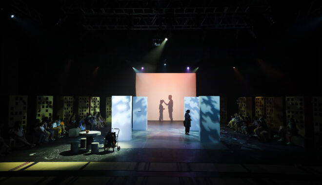 A Come Back Home production photo. A dimly lit stage foregrounds a square backdrop. A silhouette of an adult facing a child is cast in shadows on the backdrop. Photo: Studio Znke, courtesy of Esplanade - Theatres on the Bay, Singapore