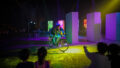 A Come Back Home production photo. A performer illuminated under soft overhead lighting rides a bicycle on stage. An audience faces the performer. The performer rides between skyscraper buildings made from cardboard. Photo: Studio Znke, courtesy of Esplanade - Theatres on the Bay, Singapore