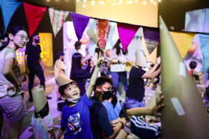 A Come Back Home production photo. A child wearing a paper hat joyfully points to colourful fabric bunting hanging above them. Other children and families create and play in the background. They are on a large stage, illuminated with bright theatrical lighting. Photo: Studio Znke, courtesy of Esplanade - Theatres on the Bay, Singapore