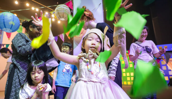 A Come Back Home production photo. A child wearing a white dress throws squares of green paper in front of them. The paper is in the air and is falling towards the ground. Photo: Studio Znke, courtesy of Esplanade - Theatres on the Bay, Singapore