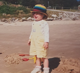 A child is standing on a sandy beach. They are wearing a colourful hat with yellow overalls and a white long-sleeved shirt underneath.