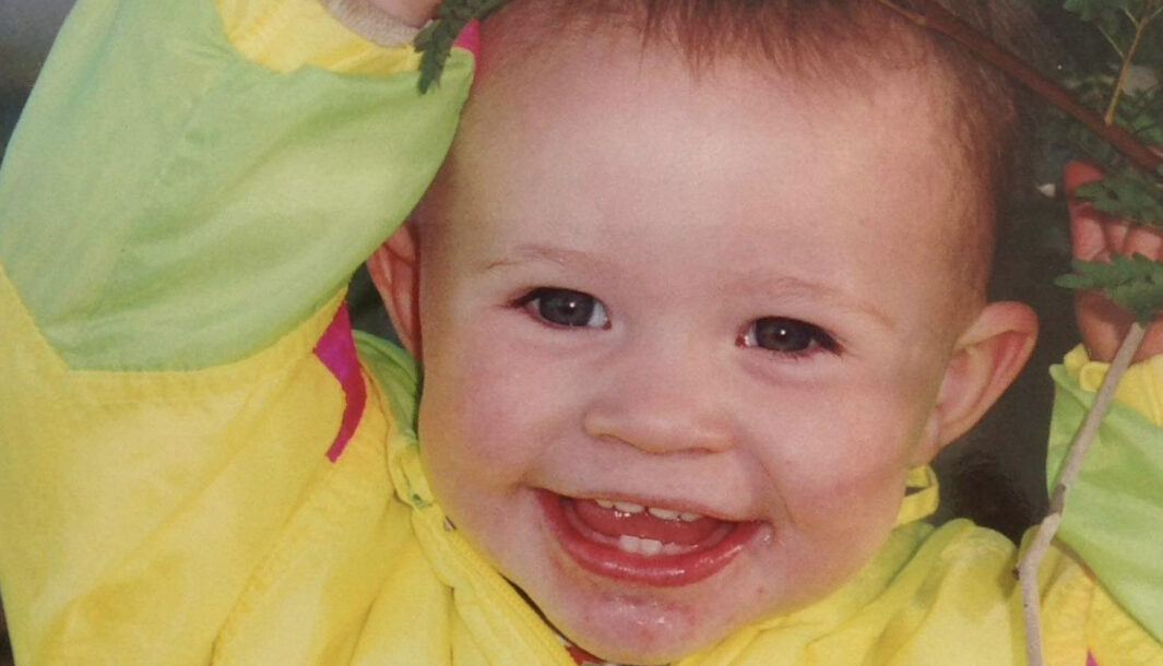 A baby is smiling. Their mouth is wide open. They have short brown hair and are wearing a yellow coat.