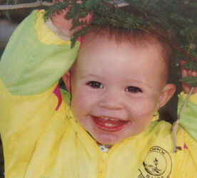 A baby is smiling. Their mouth is wide open. They have short brown hair and are wearing a yellow coat.