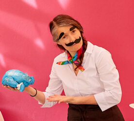 A performer stands in front of a pink background. They hold a blue, spherical object in their right hand and gesture to it with their left hand. They have glued bold and black eyebrows and a bold and black moustache on their face.