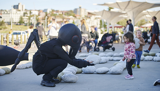 An Ants production photo. A Polyglot artist in an intricate black Ant costume crouches on a paved surface, offering a giant crumb to a small child. They are surrounded by a formation of crumbs on the ground, and children and families are visible in the background. Photographer: Lucy Parakhina, courtesy of Bondi Festival.