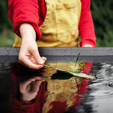 A FLOAT promo image. A child in a red jumper and yellow overalls carefully places a handmade leaf boat into a pool of water. Their hand, torso, and the boat can be seen in the reflection. Photographer: Darren Gill.