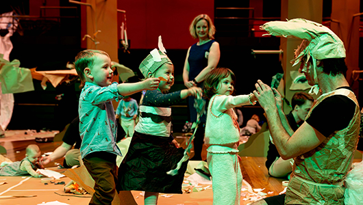 A Paper Planet production photo. A Polyglot artist in a handmade brown paper costume engages with three small children who all reach towards them. An adult in the distance looks on. They are indoors among a forest of tall cardboard trees, illuminated with bright theatrical lighting. Other children and families create and play in the space. Photo: Katje Ford, courtesy of Sydney Opera House
