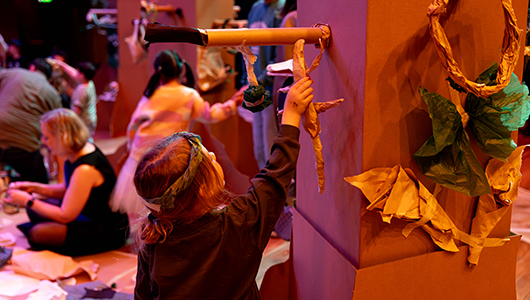 A Paper Planet production photo. A child carefully places a paper creature in a cardboard tree. Other children and families create and play in the background. They are in a darkened space, illuminated with theatrical lighting. Photo: Katje Ford, courtesy of Sydney Opera House