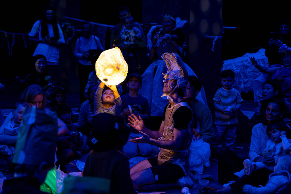 A Paper Planet production photo. Children and families are gathered closely. They are in a darkened space, illuminated with deep blue theatrical 'nighttime' light. A child reaches up to touch the moon, which illuminates them and the Polyglot artist next to them. Photo: Katje Ford, courtesy of Sydney Opera House