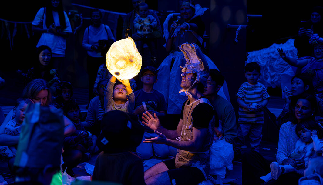 A Paper Planet production photo. Children and families are gathered closely. They are in a darkened space, illuminated with deep blue theatrical 'nighttime' light. A child reaches up to touch the moon, which illuminates them and the Polyglot artist next to them. Photo: Katje Ford, courtesy of Sydney Opera House