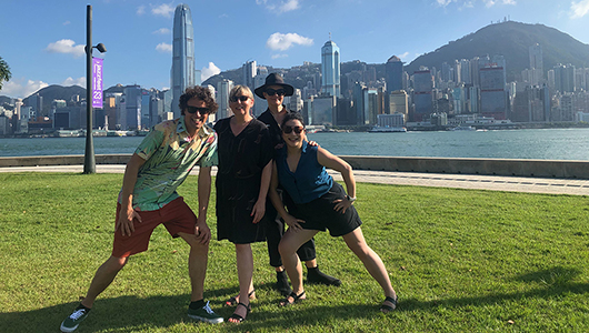 A photo of the Polyglot creative team (four people) in Hong Kong on a sunny day. They are on a green lawn next to a bay, with the city skyline and mountains in the background.