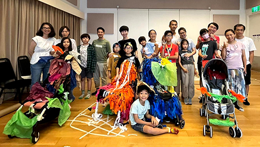 A photo of the families who participated in a Pram People development workshop in Hong Kong. They are smiling at the camera, alongside prams decorated with brightly coloured tissue paper and masking tape.