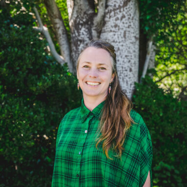 A photo of Cat Sewell, Polyglot's Artistic Director and co-CEO. She has long hair, and wears a green plaid shirt. She stands in front of a large tree and bank of green shrubbery, smiling at the camera. Photographer: Theresa Harrison