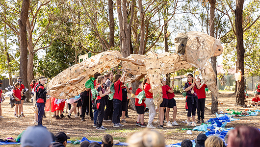 A Totems production photo. A group of students hold up an enormous handmade paper creature. They are outdoors, among trees. Gathered family and friends look on. Photographer: Alicia Fox