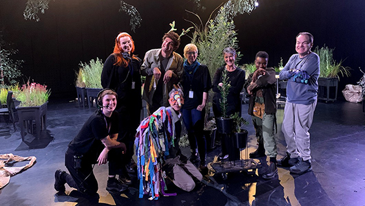 A photo of the When the World Turns team in the performance space, smiling at the camera. They are surrounded by plants in tubs and stands, branches suspended from the ceiling, and large pieces of wrinkled brown paper, illuminated by bright theatrical lighting.
