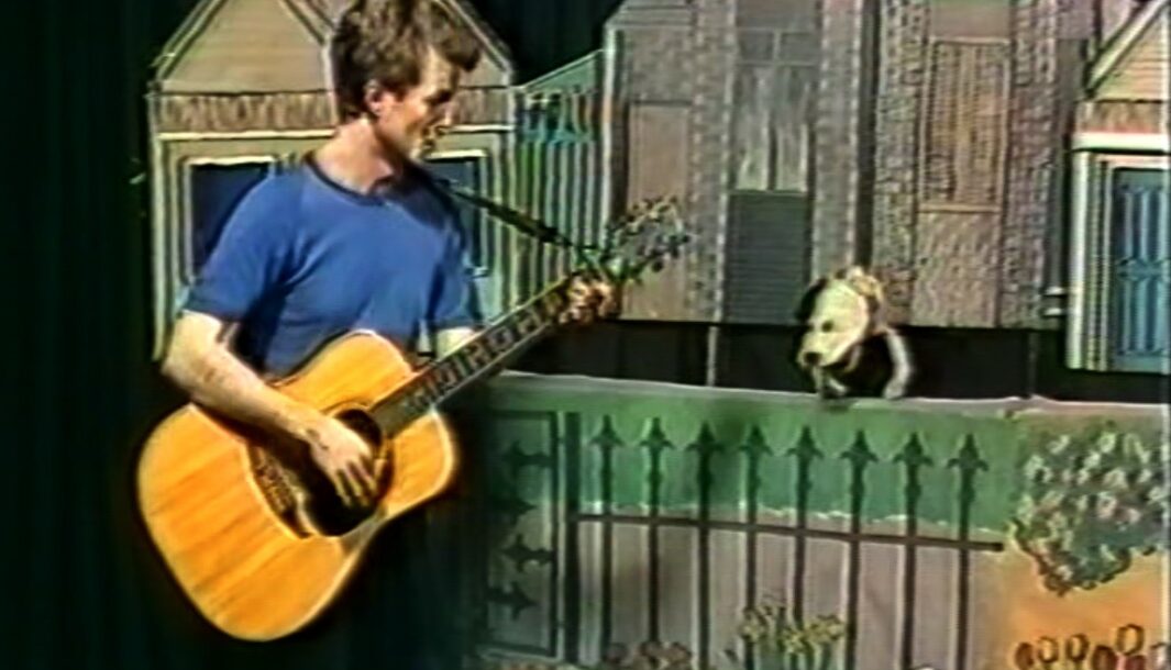 A screengrab from the 'Our Street' production video in 1987. A man in a blue shirt plays a guitar and sings to a dog puppet who rests on top of a barrier painted as a metal rod fence, with painted flowers underneath to resemble a park fence. Behind the fence and the dog is a painted row of wooden houses.