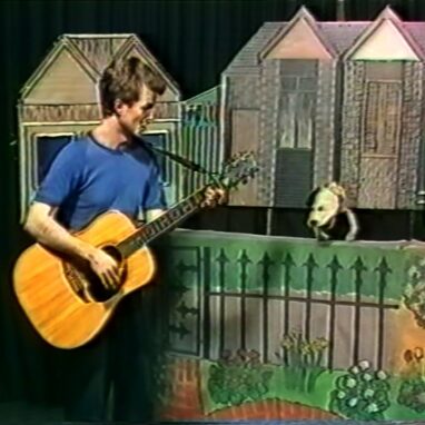 A screengrab from the 'Our Street' production video in 1987. A man in a blue shirt plays a guitar and sings to a dog puppet who rests on top of a barrier painted as a metal rod fence, with painted flowers underneath to resemble a park fence. Behind the fence and the dog is a painted row of wooden houses.