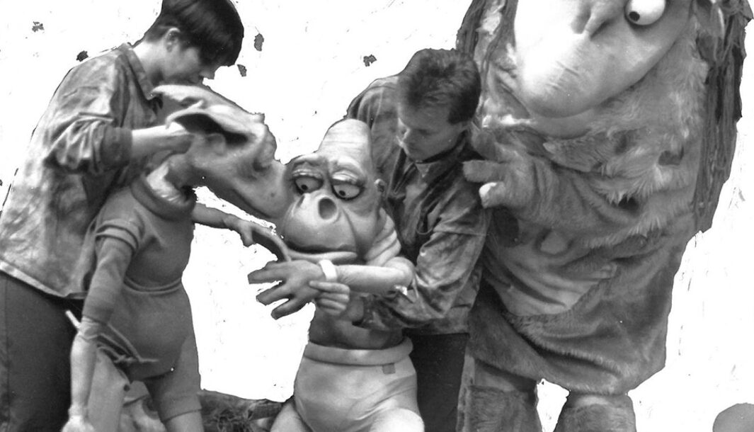 A 'Waste of Space' production photo. Two men hold two life-size puppets who have large eyes and ears. The man on the right is standing in front of a larger, furry puppet twice his size.