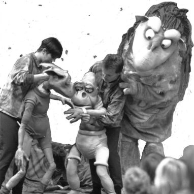 A 'Waste of Space' production photo. Two men hold two life-size puppets who have large eyes and ears. The man on the right is standing in front of a larger, furry puppet twice his size.