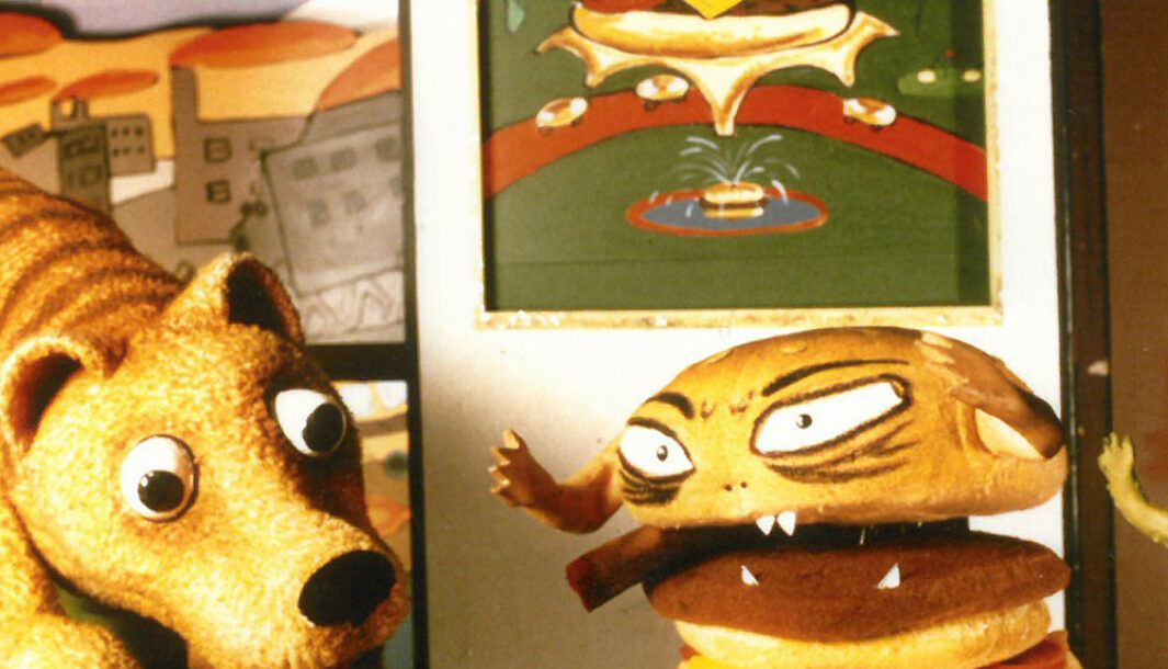 A 'Not the End of the World' production photo. A Tasmanian Tiger puppet interacts with a hamburger puppet. Behind them is a picture of a floating hamburger with a flag pitched on top.