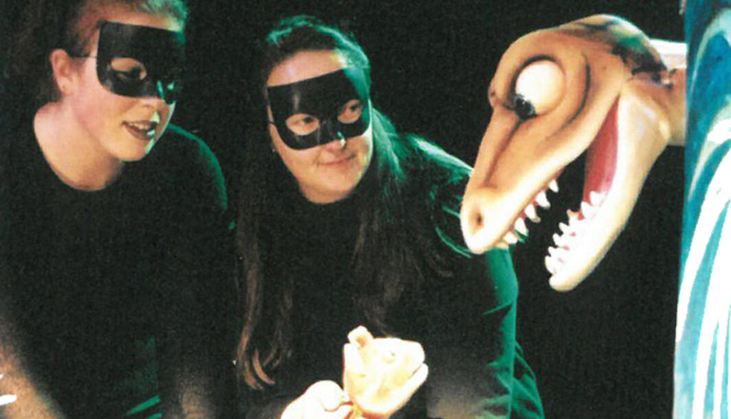 An Almost a Dinosaur production photo. Two performers wear black masks look at a dinosaur puppet that has its mouth wide open, facing sideways.